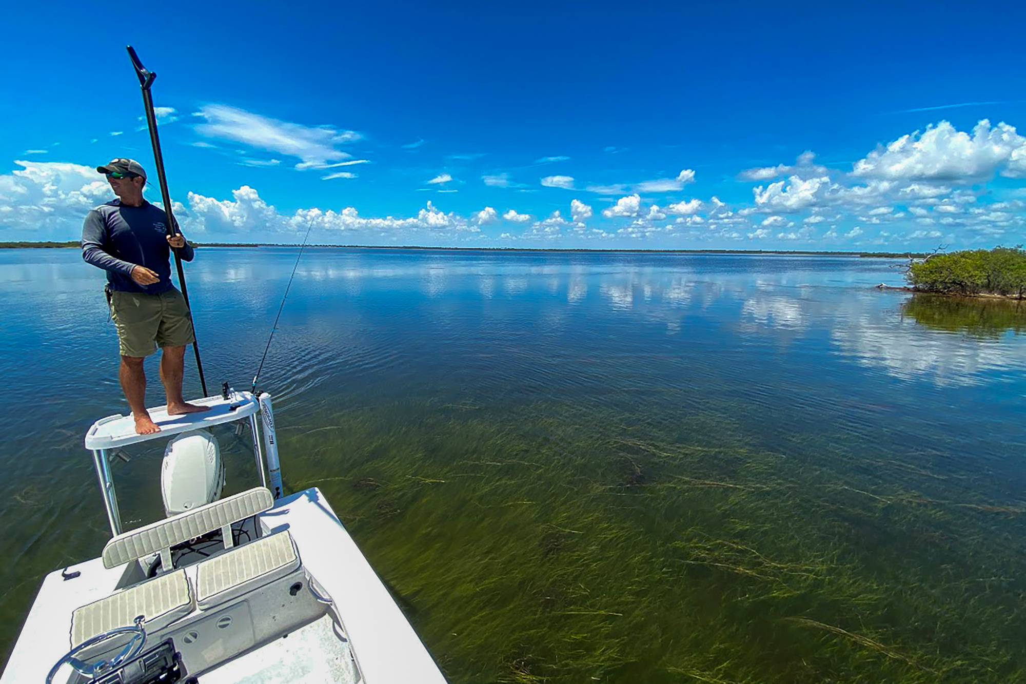 A fisherman standing on the poling platform of his skiff in the Indian River Lagoon on a clear-water day. Recovering seagrass beds visible under the surface, mangroves along the shoreline, blue sky reflected in calm water.