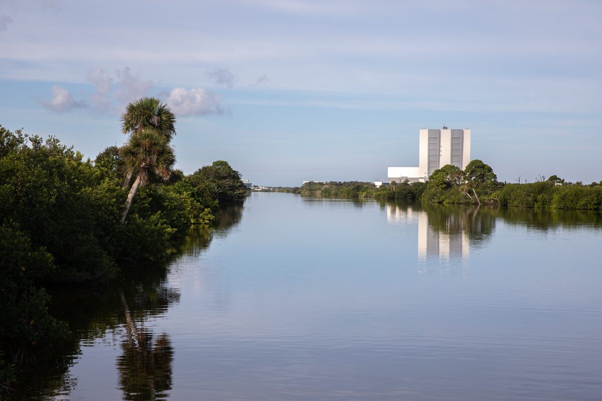 Indian River Lagoon water and shoreline with Kennedy Space Center's Vehicle Assembly Building visible in the distance under a bright sky.